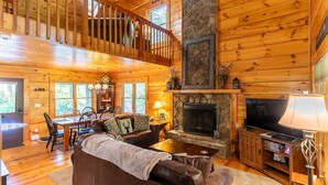 A spacious log cabin interior with a dining area, a fireplace, a television, and a balcony.
