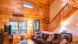 A cozy cabin interior with a wooden interior, a leather sofa, a coffee table, a bookshelf, and a staircase.