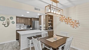 A modern kitchen with white cabinets, a central island, and a dining area with a wooden table and white chairs. - 103 Beach Level, Beautifully Decorated, Ground Floor with FREE Beach Service (Destin)