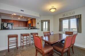 A dining area with a large wooden table and six chairs, a kitchen with wooden cabinets and appliances, and a view of the beach through large windows. - Spacious oceanfront, corner unit + Free Attraction Tickets! (North Myrtle Beach)