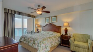Habitación de hotel con cama, dos mesitas de noche con lámparas, una silla, ventilador de techo y vista al mar.