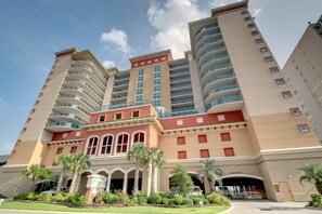 A multi-story hotel with a central red and beige facade, multiple balconies, and a covered entrance area with palm trees. - Spacious oceanfront, corner unit + Free Attraction Tickets! (North Myrtle Beach)