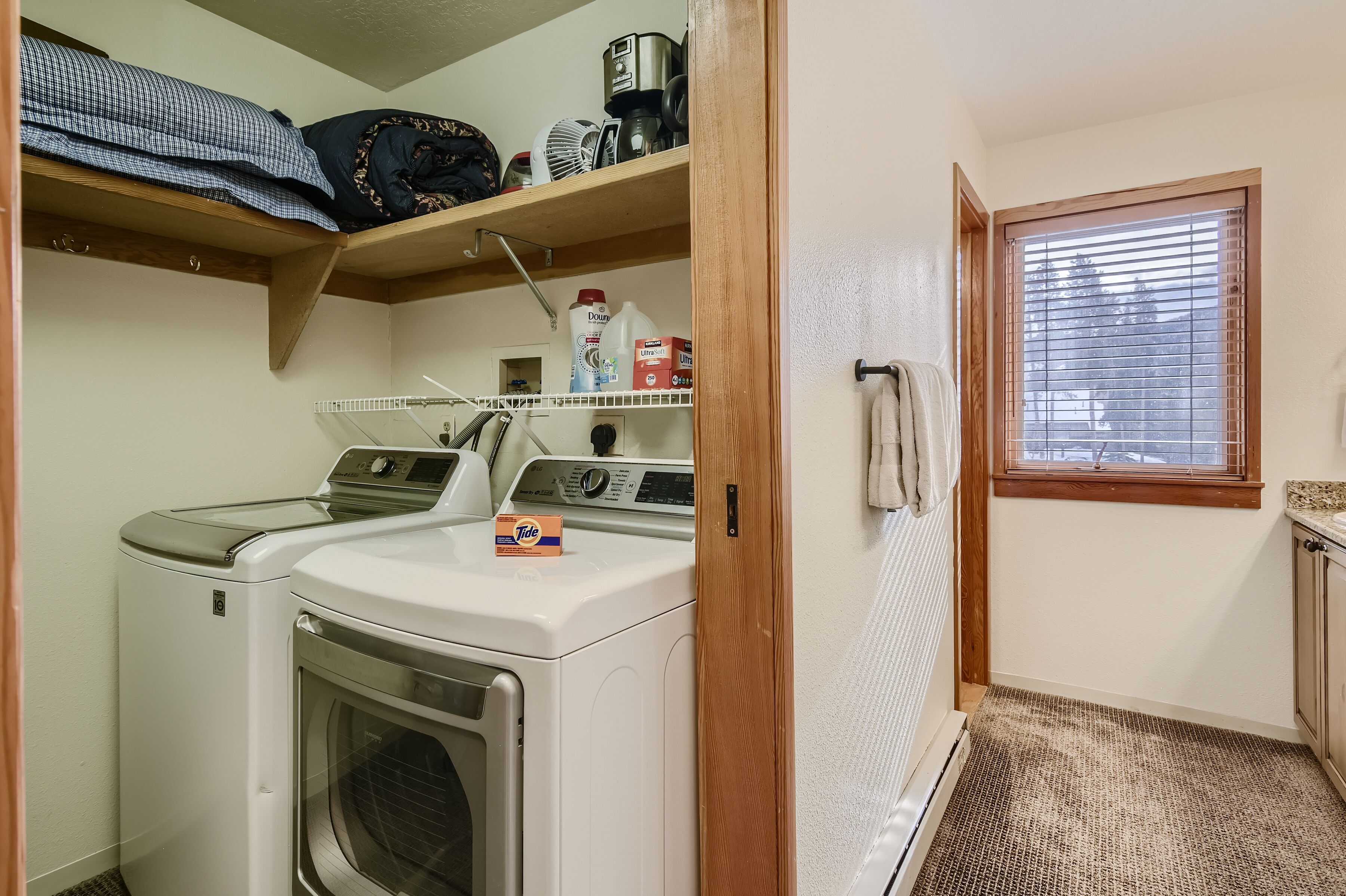 Une buanderie équipée d’un lave-linge et d’un sèche-linge à chargement frontal, d’une étagère contenant divers objets et d’une fenêtre avec des stores.