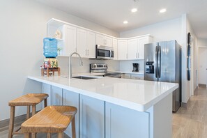 A modern kitchen with white cabinetry, stainless steel appliances, a large island with stools, and a water dispenser.