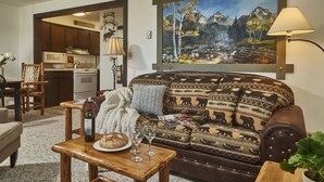 A cozy living room with a sofa, a coffee table with a loaf of bread and wine glasses, a painting of a mountain landscape, and a kitchen in the background. - JHRL - Tensleep B14, Cozy condo with skier shuttle access (Teton Village)