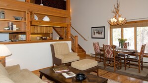 Un salon confortable avec un canapé, une table basse sur laquelle sont posés des livres, un coin repas avec des chaises et une table, et un escalier menant à un étage supérieur.