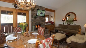 Un salon chaleureux avec une cheminée en pierre, une table à manger en bois dressée pour quatre personnes et un canapé confortable.