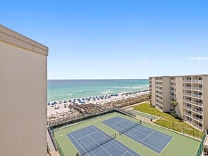 Uma quadra de tênis com vista para a praia, uma piscina e um edifício com varandas.
