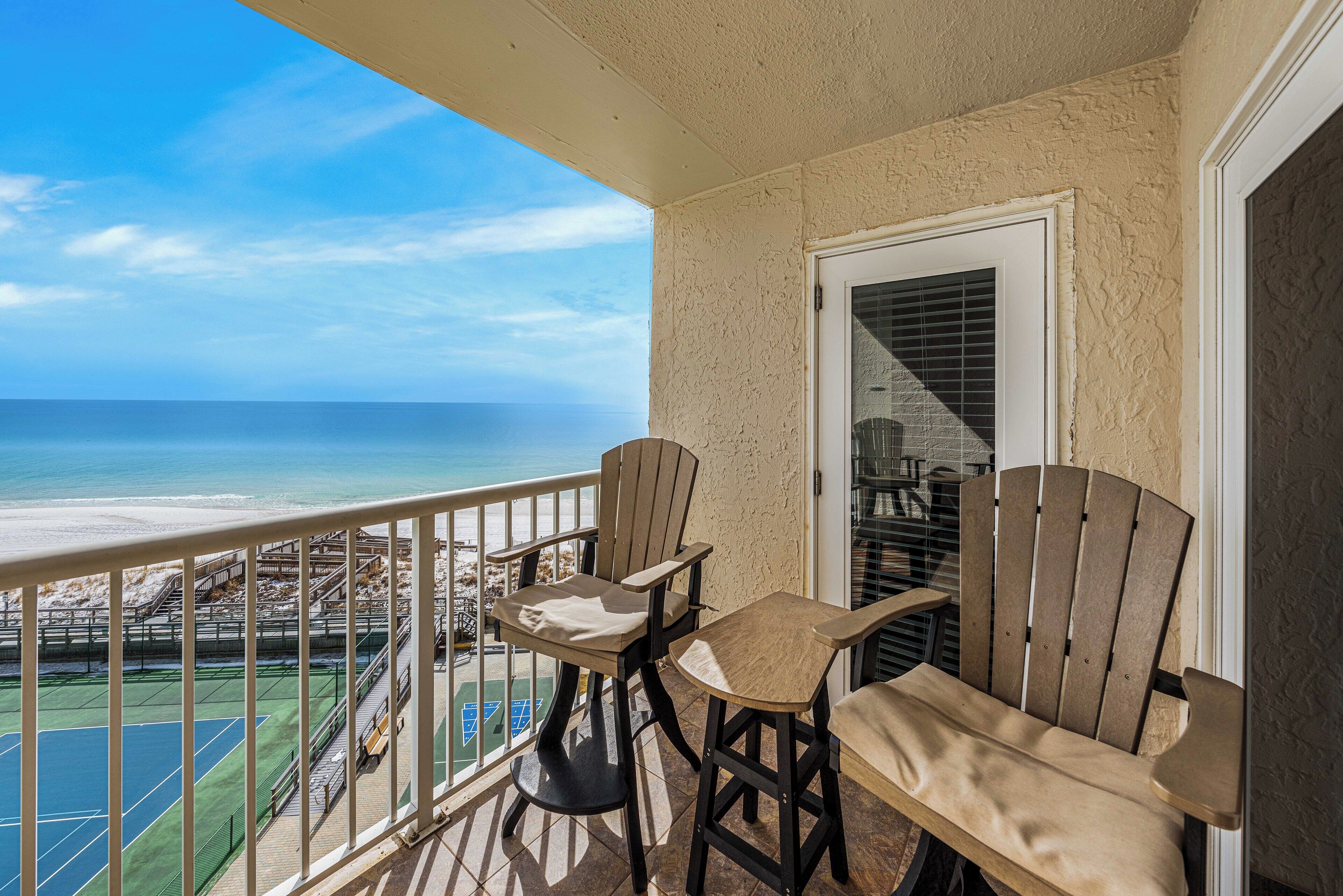 A balcony with two Adirondack chairs and a table, overlooking a beach and ocean.