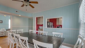 Un comedor con una mesa de madera y sillas blancas, un ventilador de techo y vista hacia una cocina con paredes rojas.