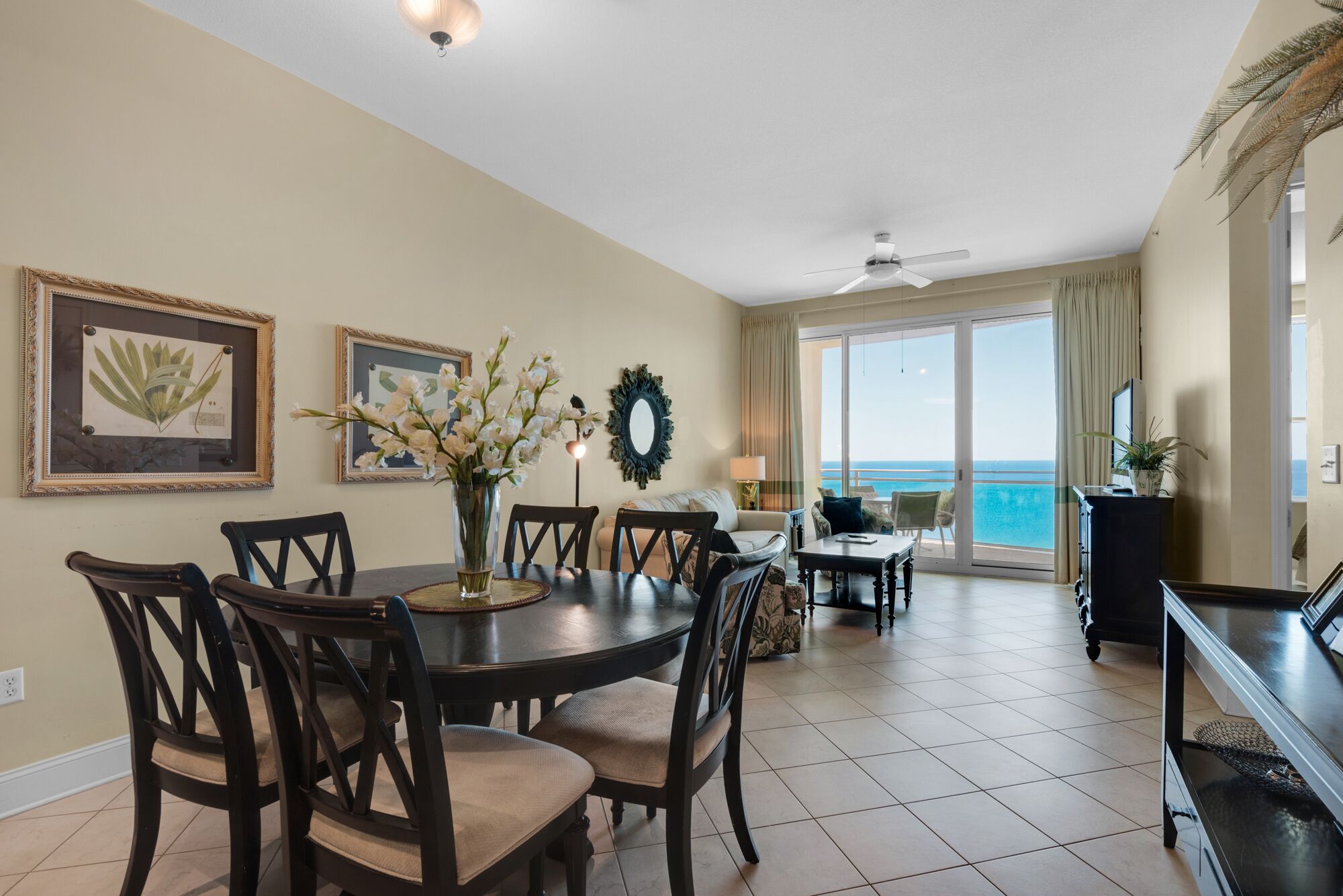 A dining area with a round table, chairs, and a vase with flowers. There are framed pictures on the wall, a ceiling fan, and a view of the ocean through large windows.