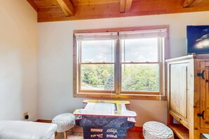 A room with a wooden ceiling, a window with a view of trees, a table with a Pac-Man game, and a cabinet.