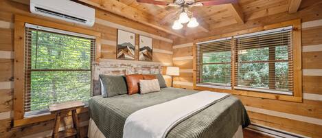 A log cabin bedroom with a bed, a nightstand, a ceiling fan, and two windows with blinds.