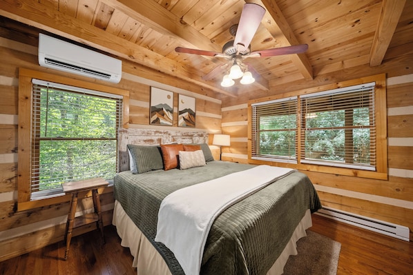 A log cabin bedroom with a bed, a nightstand, a ceiling fan, and two windows with blinds.