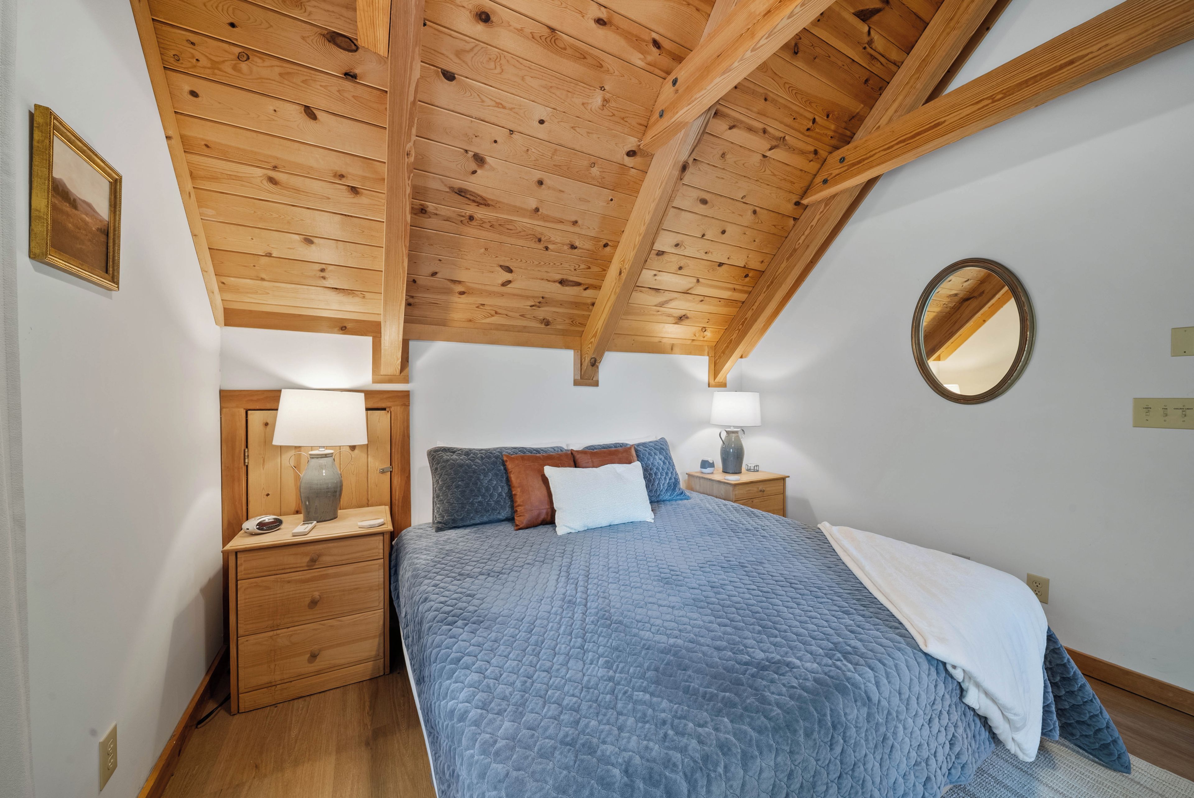 A bedroom with a wooden ceiling, a bed with a quilted blue cover, bedside lamps, a bedside table, and a mirror on the wall.