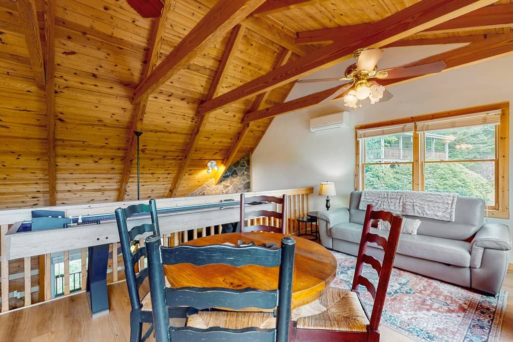 A wooden ceiling with exposed beams, a round wooden table with chairs, a sofa, and a window with a view of greenery.