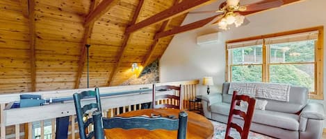 A wooden ceiling with exposed beams, a round wooden table with chairs, a sofa, and a window with a view of greenery.