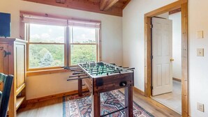 A room with a wooden ceiling, a window with a view of greenery, and a foosball table. - A Sunrise Summit - 4BR, 3BA - Hot Tub, Pets (Seven Devils)