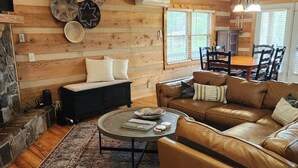 A cozy living room with a stone fireplace, leather sofas, a dining table, and a large window. - A Sunrise Summit - 4BR, 3BA - Hot Tub, Pets (Seven Devils)