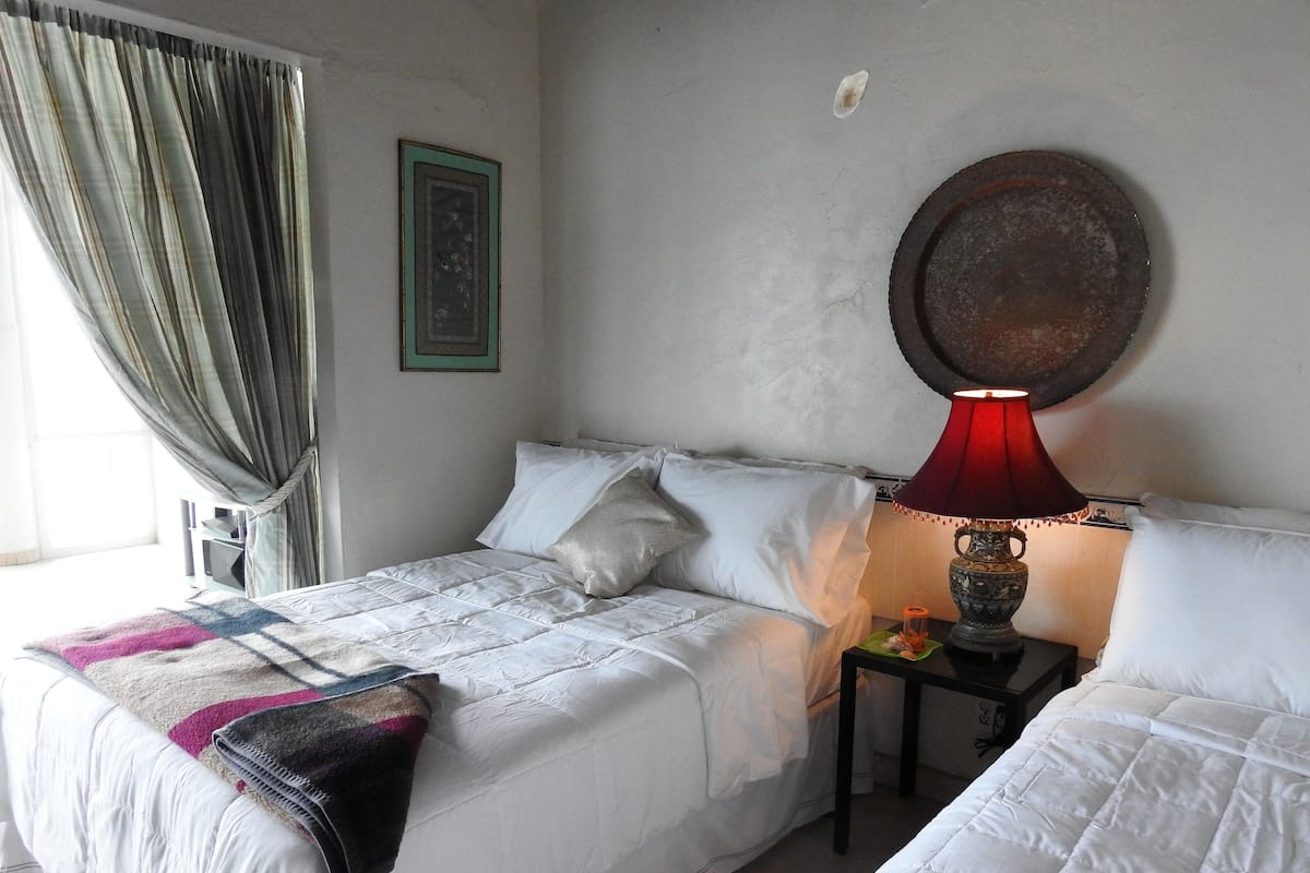 Une chambre avec deux lits, une lampe rouge, un tableau encadré et une fenêtre avec des rideaux.
