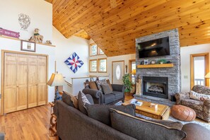 A cozy living room with a stone fireplace, wooden ceiling, and a large TV mounted above it.