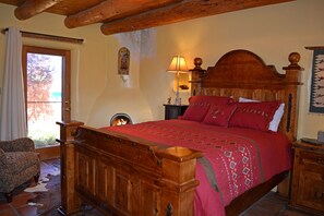 Een houten bed met een rode sprei, een nachtkastje met een lamp, en een stoel in een kamer met een houten plafond en een raam met gordijnen.