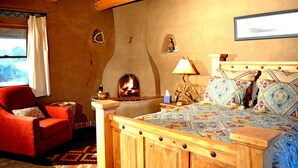 Een gezellige kamer met een houten plafond, een open haard, een bed met beddengoed met een patroon, een rode fauteuil en een klein tafeltje met een lamp.