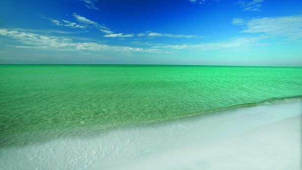 Een strand met helder turquoise water en een witte zandkust onder een blauwe lucht.
