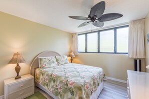 Een slaapkamer met een bed, nachtkastje, lamp, plafondventilator en een raam met gordijnen.