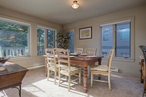 Uma área de refeições com mesa e cadeiras de madeira, janelas amplas e um vaso com planta.