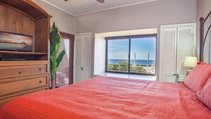 A bedroom with a large bed, a wooden dresser, a television, and a window with a view of the sea.