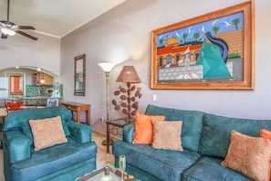 A living room with teal sofas, orange and beige cushions, a floor lamp, a painting, and a kitchen in the background.