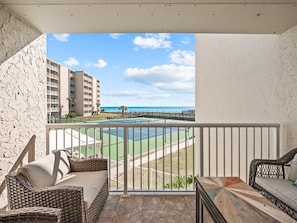 A balcony with wicker furniture, a tennis court, and a view of a beach and hotel buildings.