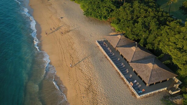 Vue aérienne d’un complexe hôtelier en bord de mer, avec un long bâtiment doté de nombreuses zones ombragées et d’une piscine.