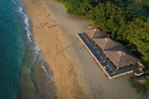 Vista aérea de un resort frente al mar, con un edificio extenso que ofrece múltiples zonas con sombra y una piscina.