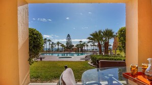A balcony with a view of a swimming pool, palm trees, and a distant building. - Princesa C-104 Barefoot Bumblebee Newly Renovated Ocean View Condo (Puerto Penasco)