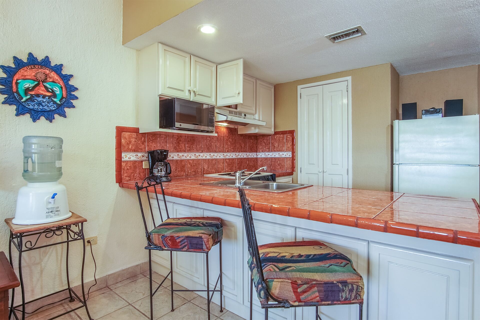 A compact kitchen with white cabinets, a stainless steel sink, and a microwave. There are two colorful stools and a water dispenser on a small table.