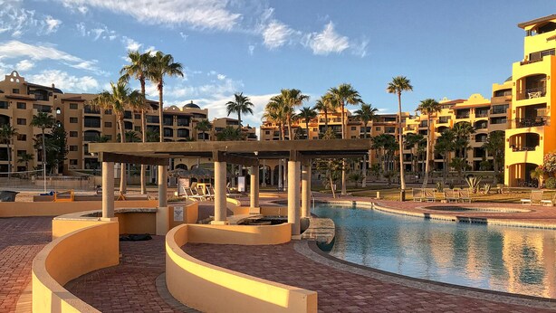 A resort pool area with a curved water feature, palm trees, and a covered seating area. - Princesa C-104 Barefoot Bumblebee Newly Renovated Ocean View Condo (Puerto Penasco)