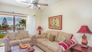 A living room with a sofa set, two side tables with lamps, a ceiling fan, and a large window with a view of a pool and palm trees. - Princesa C-104 Barefoot Bumblebee Newly Renovated Ocean View Condo (Puerto Penasco)