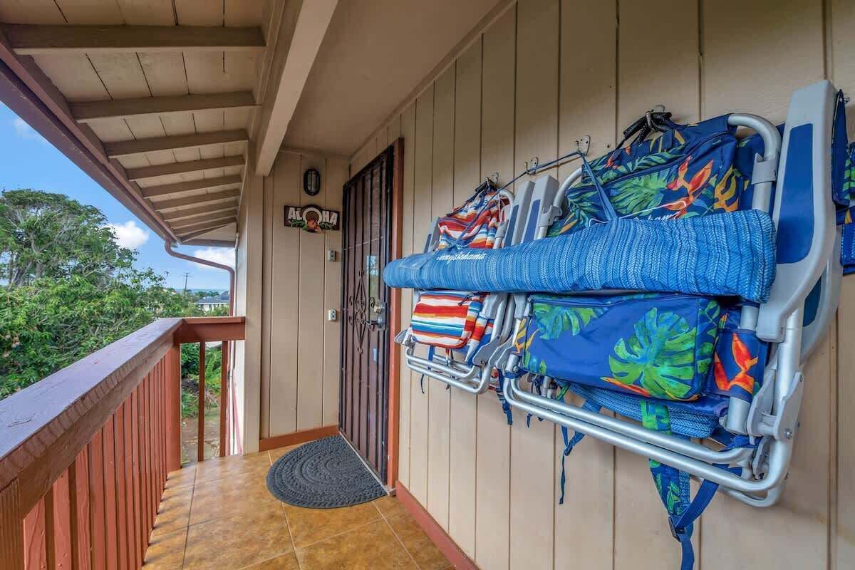 Eine überdachte Veranda mit Holzgeländer, eine Tür mit einem Schild und mehrere Strandstühle und Sonnenschirme, ordentlich an der Wand angeordnet.