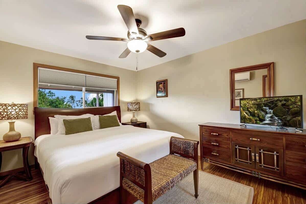 Une chambre d’hôtel avec un grand lit, un ventilateur de plafond, une télévision et une vue sur la verdure par la fenêtre.