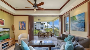 A spacious living area with a large flat-screen TV, comfortable seating, a ceiling fan, and a balcony with a view of a multi-story building and palm trees.