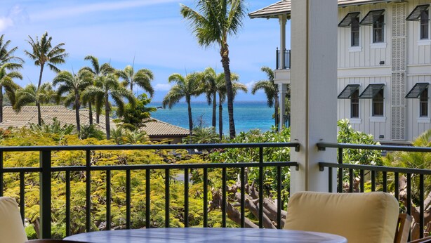 A balcony with a view of the ocean, palm trees, and a building with patterned shutters.