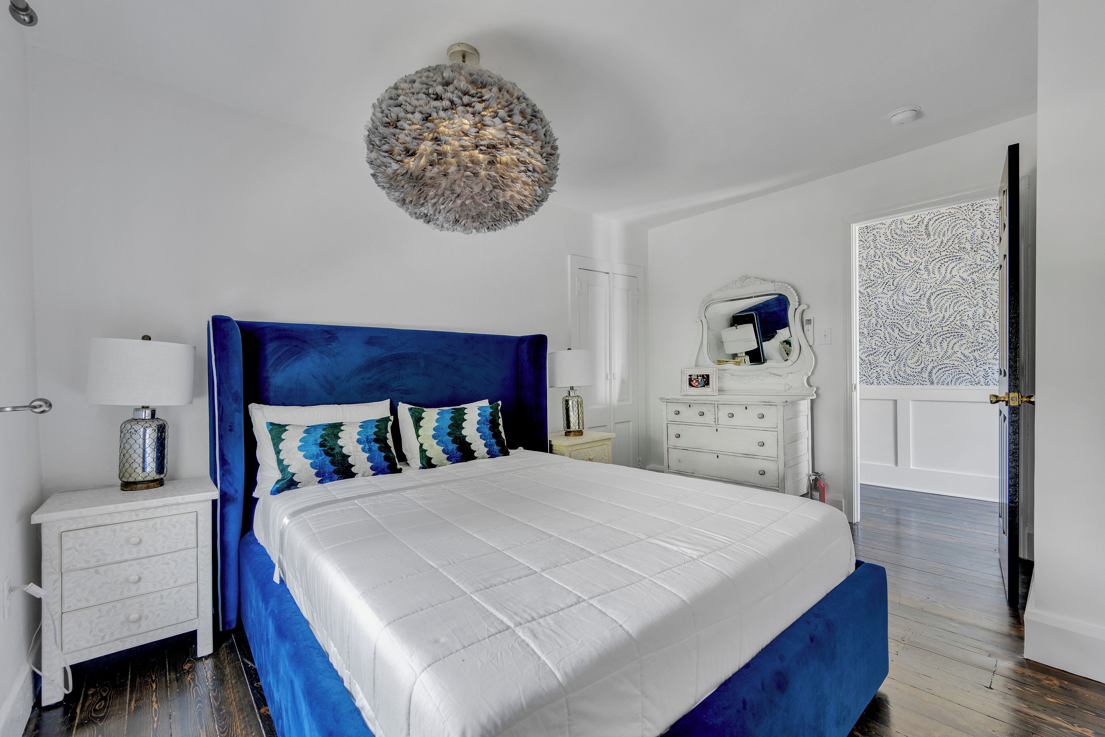 A bedroom with a large bed, white nightstands, a blue headboard, a decorative ceiling light, and a patterned door.