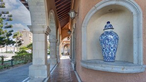 A corridor with arches and a tiled floor, leading to a building with a large blue and white vase on display.