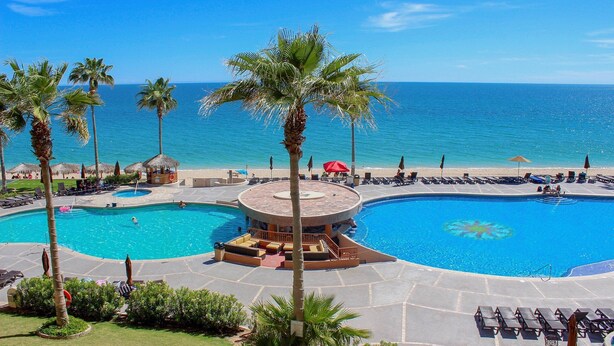 A resort pool area with two large pools, palm trees, and a beach view.