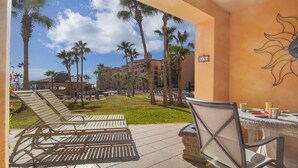 A hotel patio with lounge chairs, a dining table with chairs, and a view of the surrounding buildings and palm trees. - Princesa Resort D-104 Dream Catcher  Comfy Ground Floor Condo (Puerto Penasco)