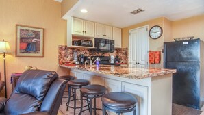 A kitchen with a granite countertop, a black refrigerator, a microwave, and a clock on the wall. There are three bar stools and a painting on the wall. - Princesa Resort D-104 Dream Catcher  Comfy Ground Floor Condo (Puerto Penasco)