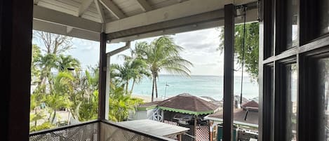 A covered balcony with a table and chairs, overlooking a beach with palm trees and a view of the ocean.
