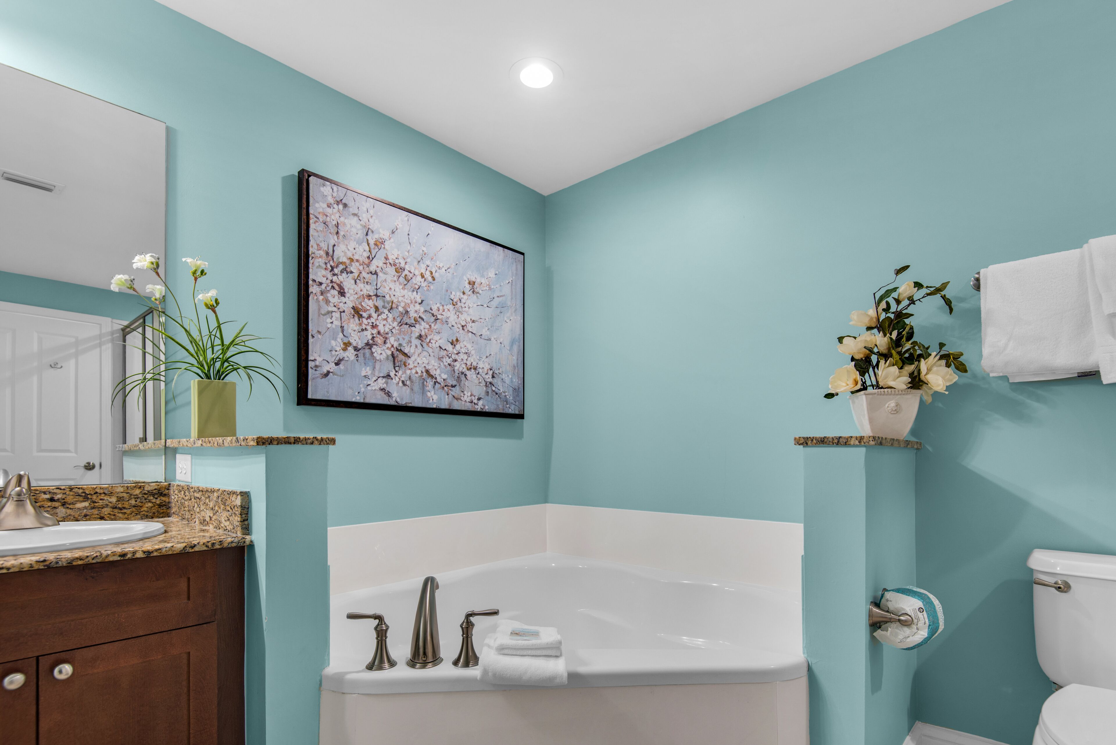 Een badkamer met een wit bad, een wastafel en een toilet. Er hangt een lijst met kunst aan de muur en op het aanrecht staat een vaas met bloemen.